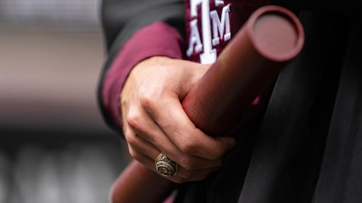 An Aggie graduate holds a diploma in the same hand wearing his Aggie ring.