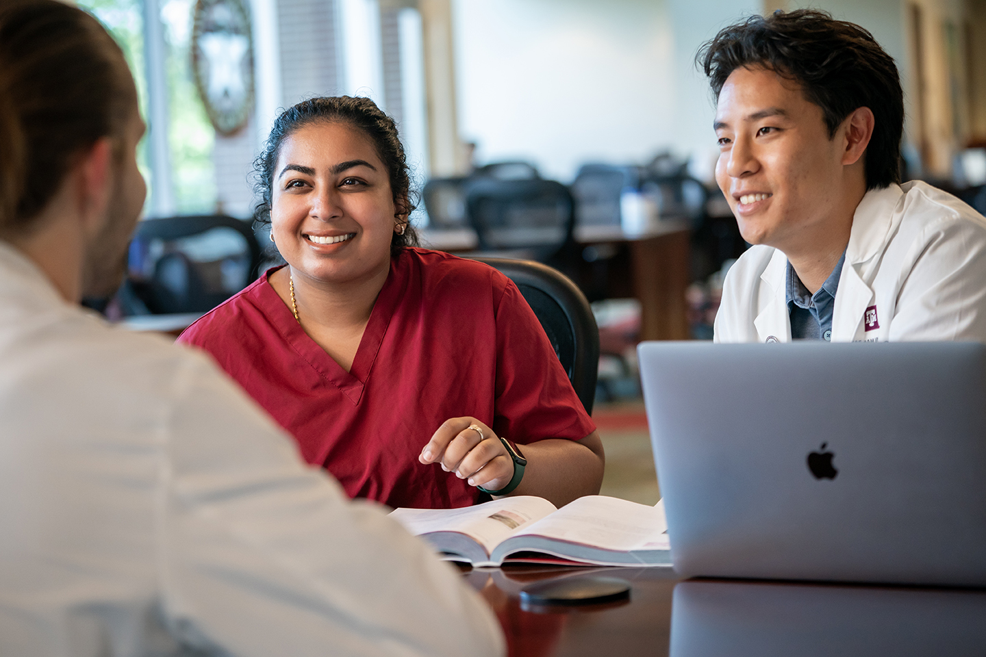 Three students having a conversation with laptop and textbook.